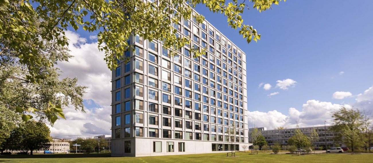 Hoog, modern gebouw met veel ramen, gefotografeerd vanaf een grasveld met bomen op de voorgrond.