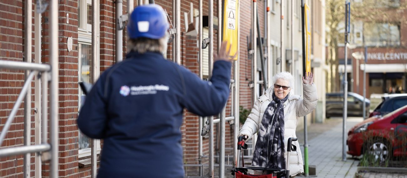 Een bewoonster zwaait vrolijk naar een medewerker van het bouwbedrijf. De medewerker met bouwhelm zwaait terug