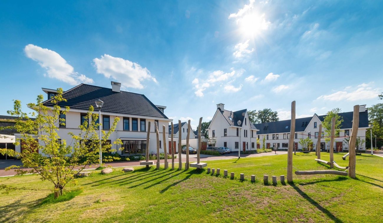 Nieuwbouwwoningen in Willemsbuiten Tilburg in karakteristieke jaren 30-stijl, met witte gevels, donkere dakpannen en een groene speelplek met houten speeltoestellen op de voorgrond onder een heldere blauwe lucht.