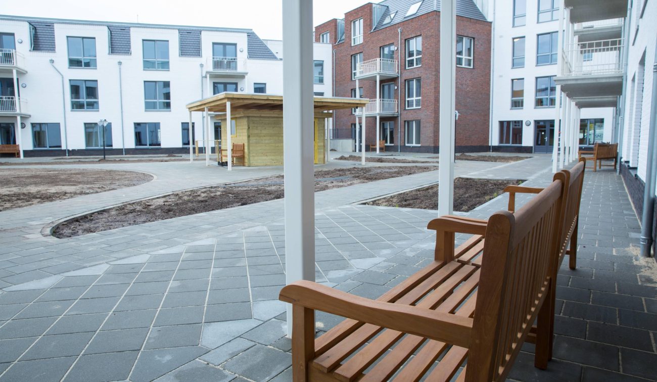 Hof te Berkel Horst – zicht vanaf overdekte galerij met houten bank op binnentuin met paden en paviljoen, appartementen met balkons