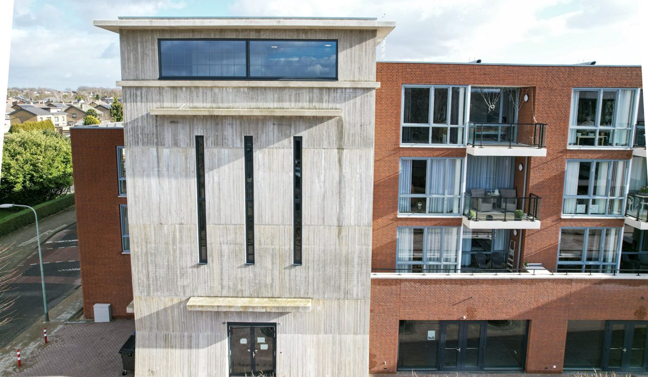 MFA Hart voor Treebeek Brunssum – betonnen torenvolume met smalle verticale ramen naast bakstenen gevel met glazen balkons