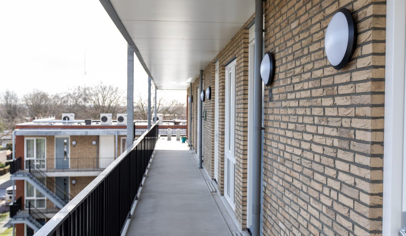 Appartementengebouw – overdekte galerij met bakstenen gevel, ronde wandarmaturen en zwart hekwerk, zicht op tegenoverliggend blok