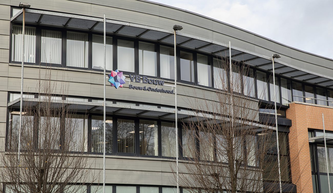 Moderne bedrijfspandgevel met groot logo en zwarte raamstroken, gezien vanaf straat met vlaggenmasten