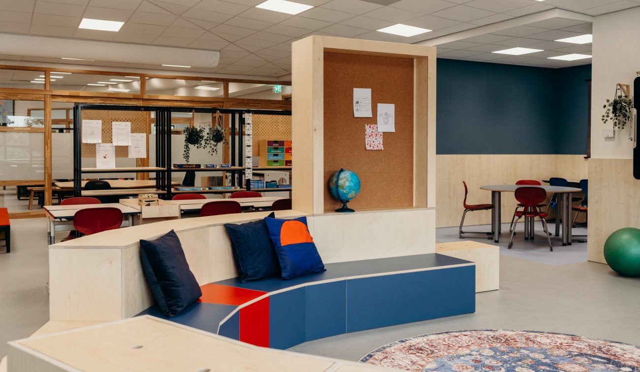Multifunctioneel leerplein met lage houten zittribune met blauwe kussens, centraal prikbord met wereldbol ervoor, groepstafels met rode stoelen op de achtergrond en glaswanden onder een systeemplafond.
