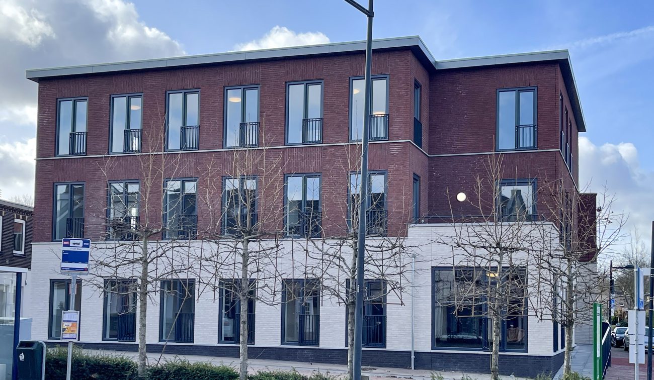 Nieuwbouw zorgcomplex Cicero Bleekstaete in Schinveld: rood­bakstenen volume op witte plint, plat dak, hoge smalle ramen met Franse balkons, straatlantaarn en bushaltebord op de voorgrond, rij leafloze bomen en blauwe lucht met wolken.