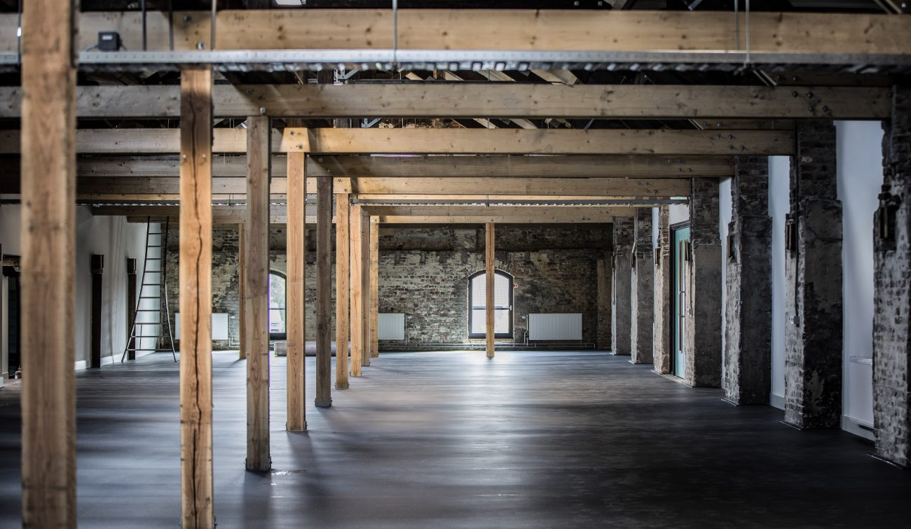 Lege industriële ruimte met houten draagbalken en kolommen, ruwe bakstenen wanden met boogvensters en een gladde donkere betonvloer; ladder links tegen de muur.