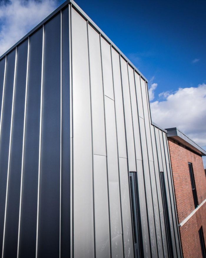 Detail van gevel met verticale metalen felsbanen in grijstinten naast een rood­bakstenen wand; smalle raampartijen, onder blauwe lucht met wolken.