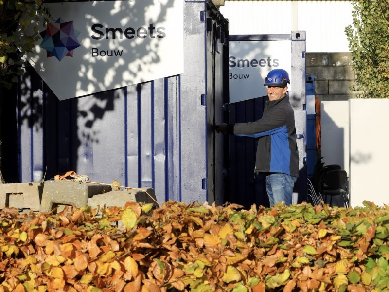 Man met een bouwhelm op pakt iets uit een opslag, met op de voorgrond een heg die verkleurd is door de herfst