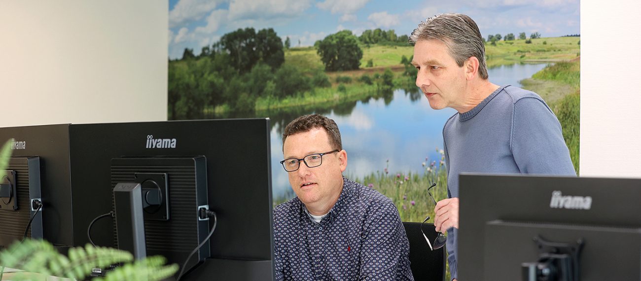 Twee collega’s overleggen achter computer­schermen in kantooromgeving met natuurbeeld op achterwand.