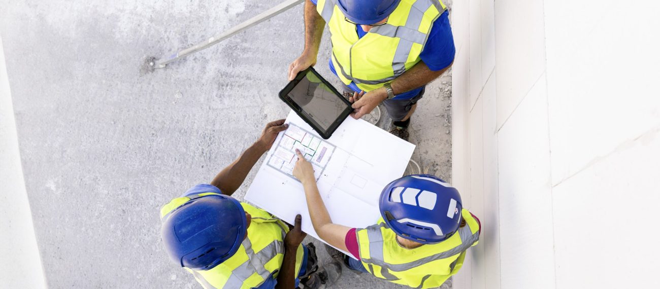 Drie bouwmedewerkers met helmen en veiligheidshesjes bekijken bouwtekeningen op een papieren plan en een tablet, gezien van bovenaf