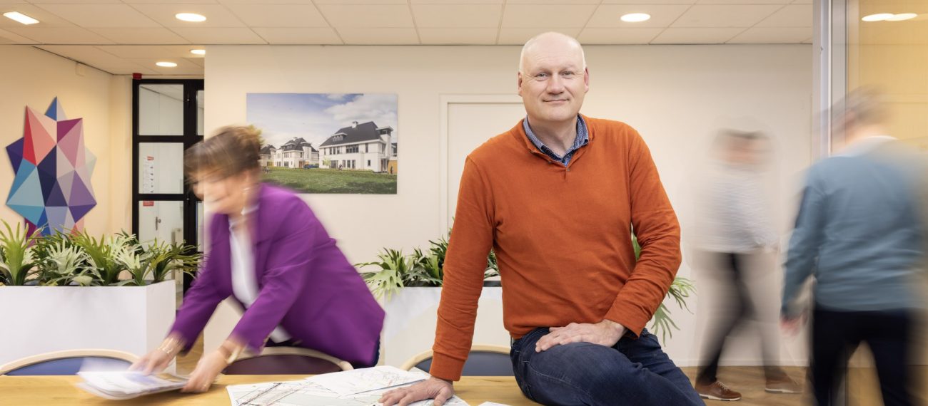 Man zit op tafel met bouwtekeningen op kantoor, collega’s bewegen op achtergrond, werksfeer vastgoedontwikkeling.