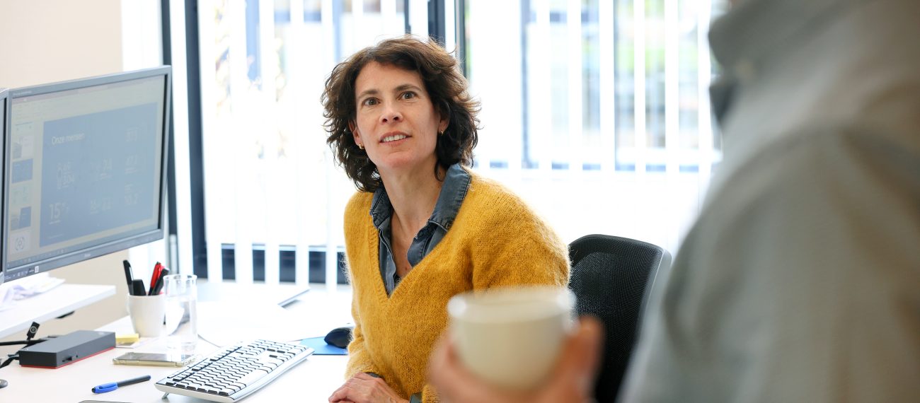 Medewerker achter bureau kijkt op naar collega met koffiemok in modern kantoor met daglicht en computer