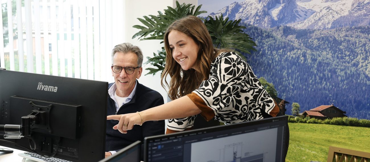 Vrolijke collega’s achter een bureau die samen naar een computerscherm kijken en iets aanwijzen, met technische tekeningen op andere schermen en een muurdecor met berglandschap op de achtergrond.