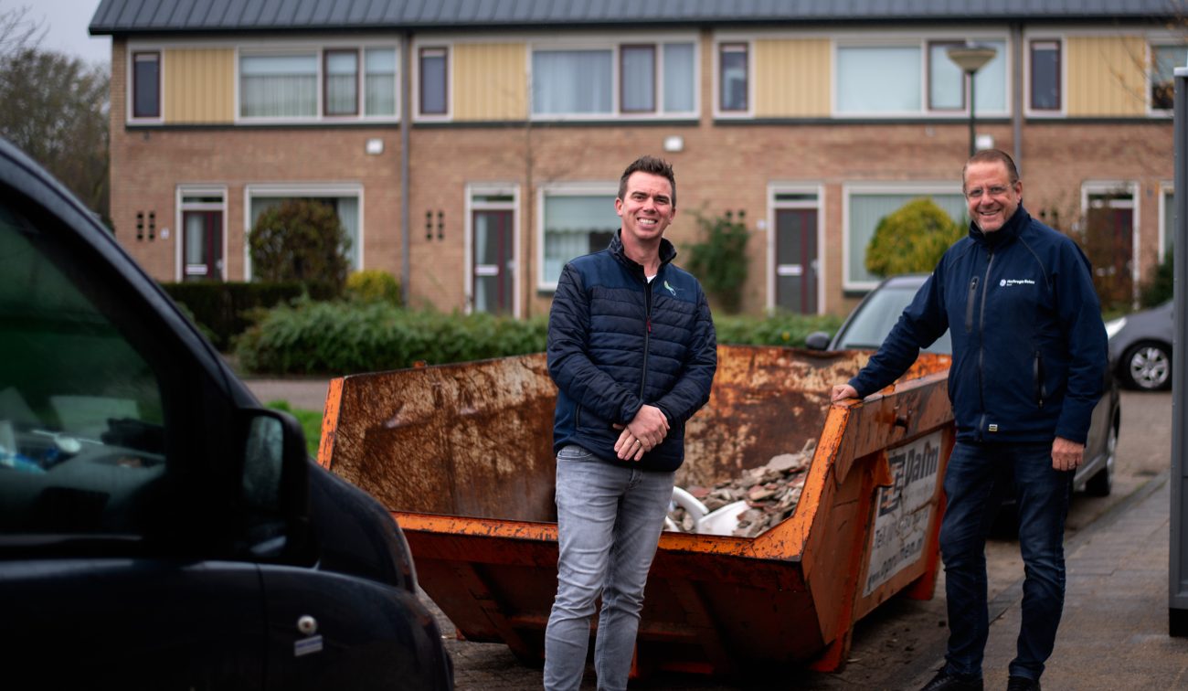 Twee mannen staan voor een afvalcontainer met op de achtergrond een rij woonhuizen met zonnepanelen