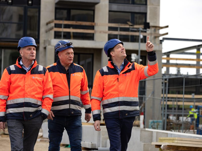 Drie mannen met bouwhelmen en oranje veiligheidsjassen lopen over een bouwplaats. Een van de mannen wijst naar boven