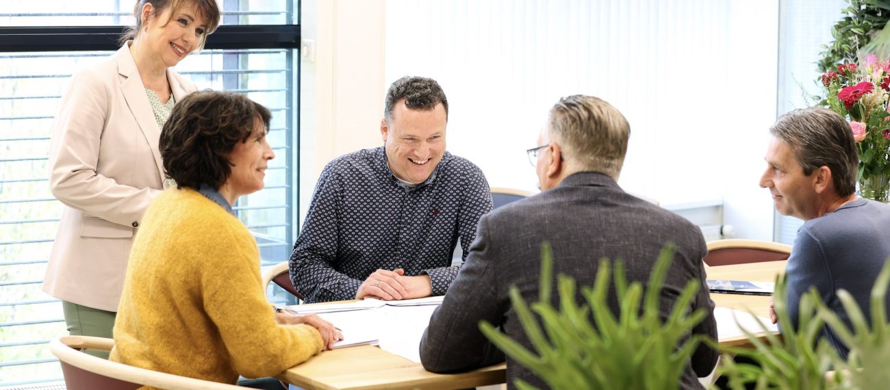 Vijf mensen zitten samen rond een tafel in een lichte vergaderruimte en praten lachend met elkaar.
