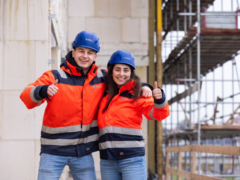 Twee mensen in oranje werkjassen en blauwe helmen staan op een bouwplaats en steken blij hun duimen omhoog.