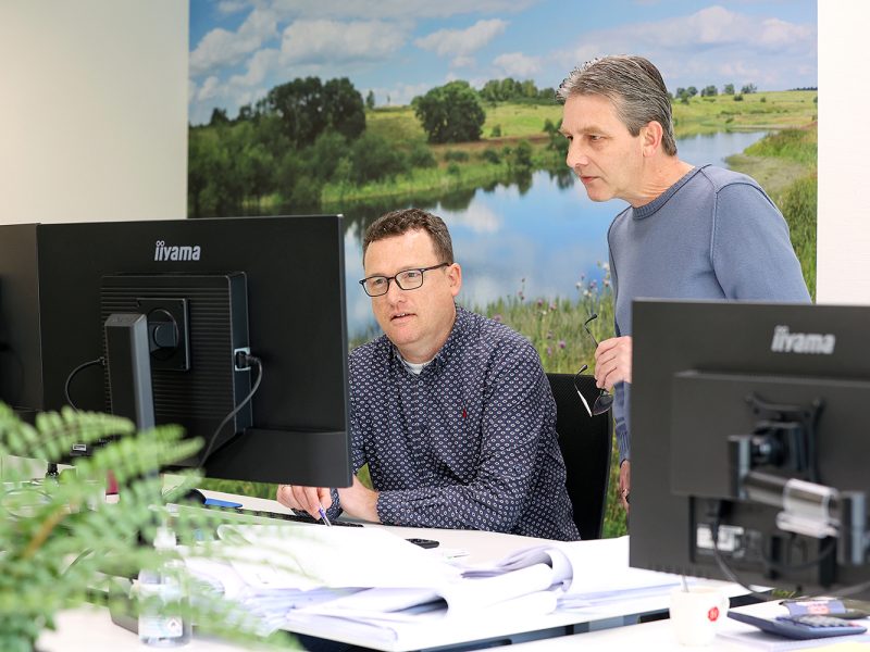 Twee collega’s overleggen achter computer­schermen in kantooromgeving met natuurbeeld op achterwand.