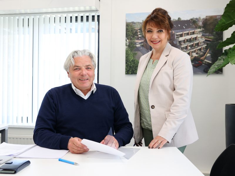 Een man en een vrouw zitten en staan aan een bureau in een lichte kantoorruimte, met documenten voor zich en een projectvisualisatie aan de muur.