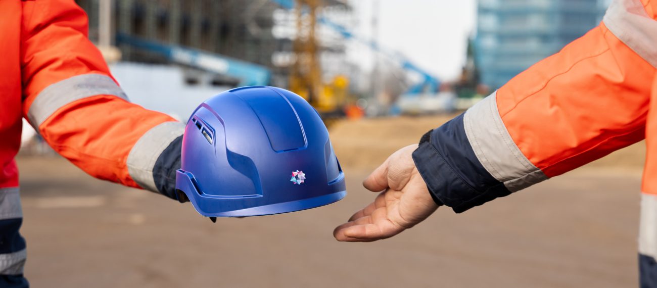 Iemand geeft een bouwhelm aan een collega, waarbij op de achtergrond twee gebouwen in de steigers staan