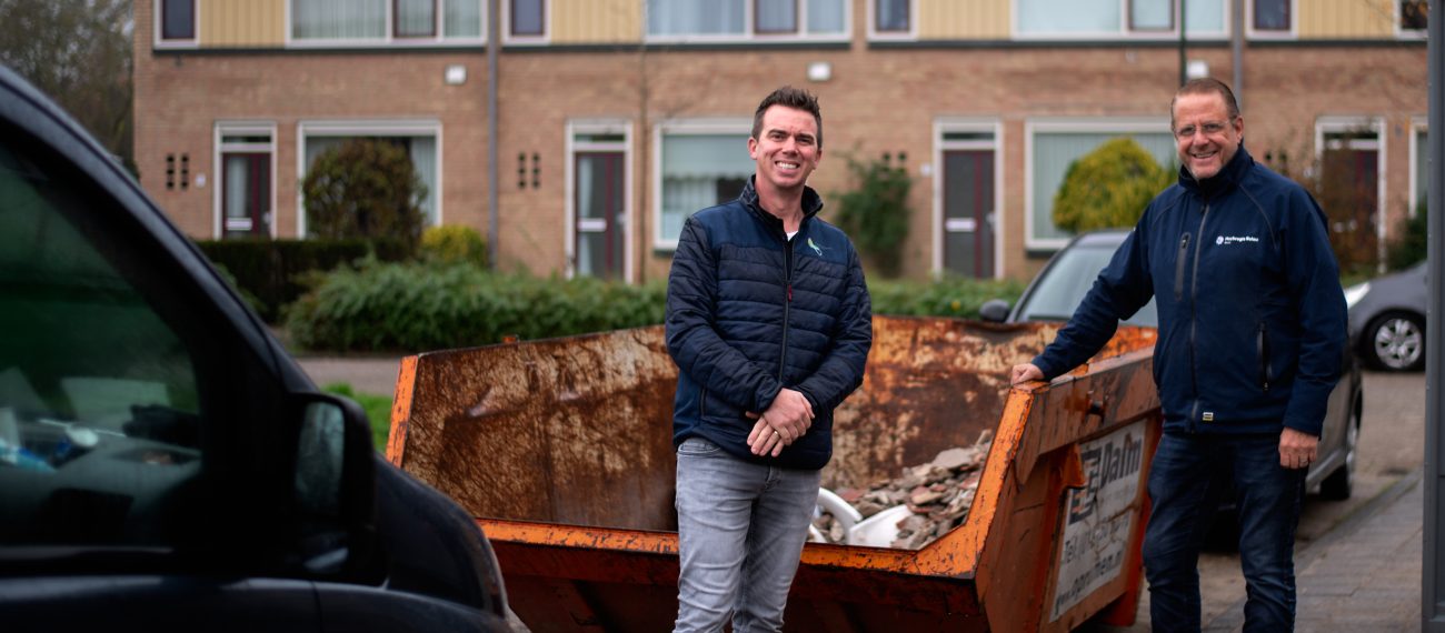 Twee mannen staan voor een afvalcontainer met op de achtergrond een rij woonhuizen met zonnepanelen