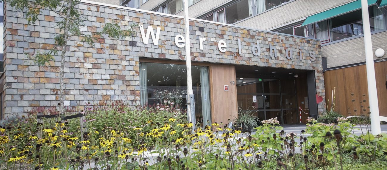 Vooraanzicht van een gebouw met de naam ‘Wereldhuis’, met een bloementuin op de voorgrond