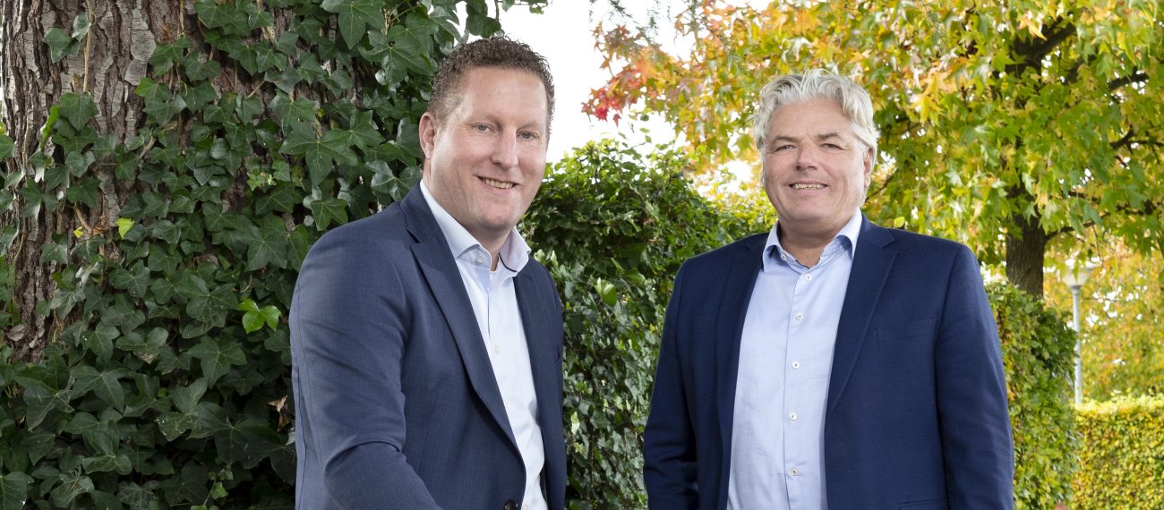 Twee mannen in pak poseren buiten bij een boom en groen struikgewas.