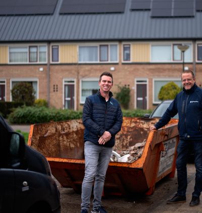 Twee mannen staan voor een afvalcontainer met op de achtergrond een rij woonhuizen met zonnepanelen