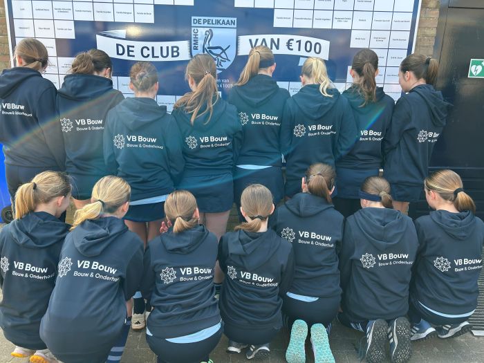 Een sportteam poseert met blauwe trainingsjacks waarop het logo van VB Bouw op de rug te zien is
