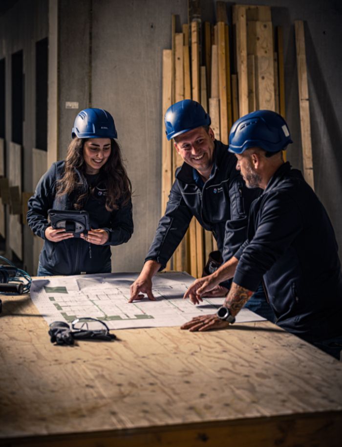 Drie mensen met blauwe bouwhelmen staan rond een tafel op een bouwplaats en bekijken bouwtekeningen; één persoon wijst op de plattegrond terwijl de anderen mee overleggen.