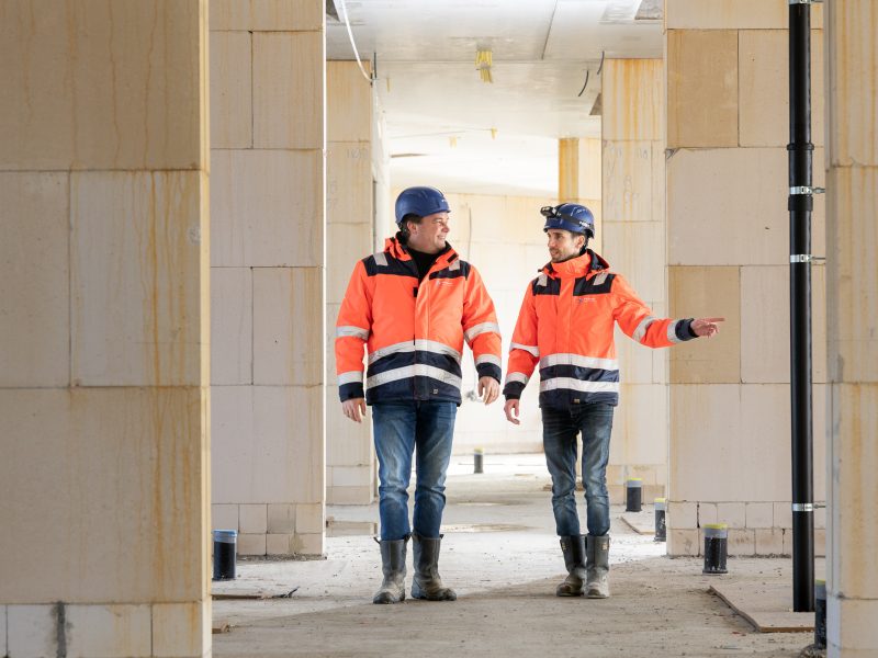 Twee collega's met veiligheidsjas en bouwhelm op lopen pratend over een bouwplaats