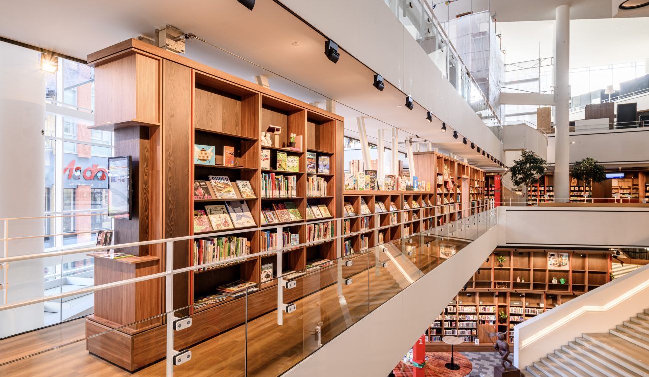 Galerijniveau met boeken en spotverlichting; glazen schermen boven de leeszone beneden.