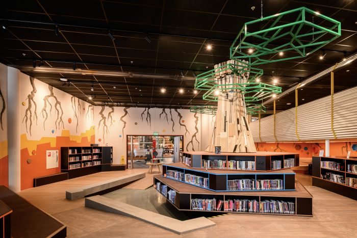 Kinderzone in bibliotheek met houten ‘boom’, felgroen frame, zitbanken en schappen vol boeken.