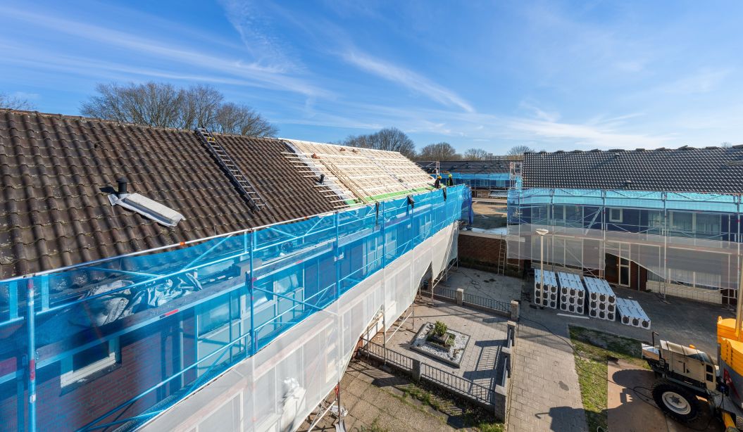 Verduurzaming Wandelbos Tilburg: woningen in steigers, dakisolatie, blauwe netten, vakmensen op dak.