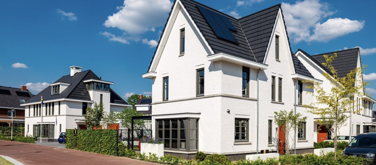 Moderne vrijstaande witte woning met donker zadeldak en zonnepanelen in een rustige woonwijk op een zonnige dag.