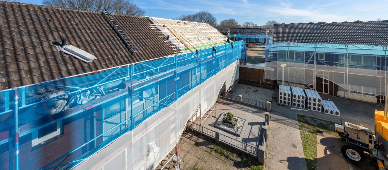 Verduurzaming Wandelbos Tilburg: woningen in steigers, dakisolatie, blauwe netten, vakmensen op dak.