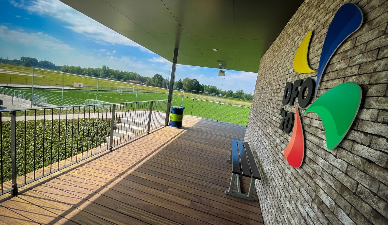 Overdekt terras van het clubgebouw DVO’20 met houten vlonder, bankje en zicht op de voetbalvelden; grote DVO’20-gevellogo op de gemetselde wand.