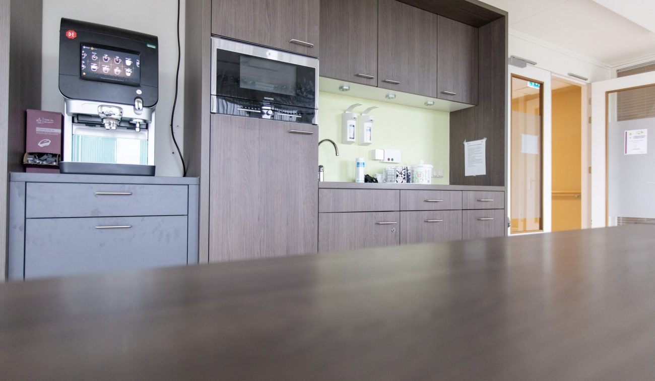 Zorgcentrum-pantry na renovatie: grijs-houtlook kastenwand met magnetron, spoelbak en lichtgeel achterwand; links professionele koffiemachine, rechts doorgang naar gang.