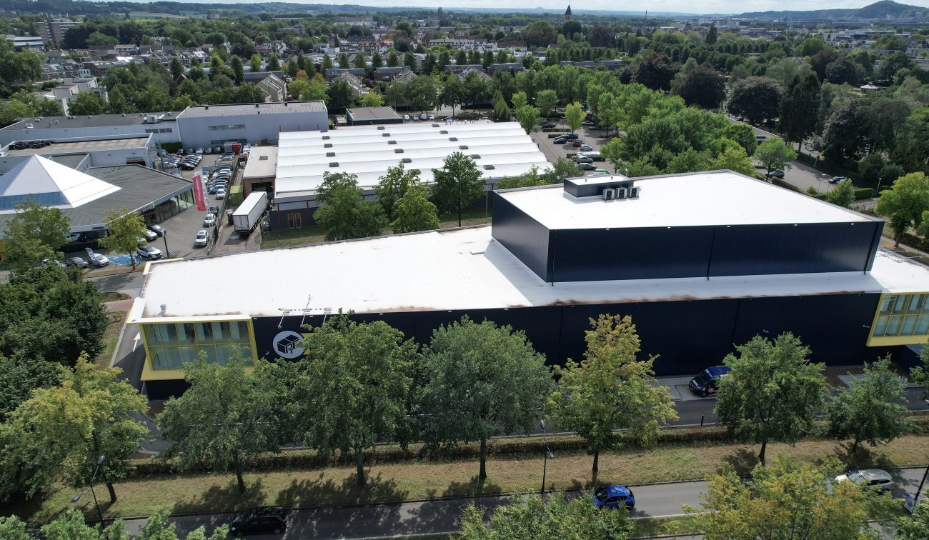 Schuine dronefoto van ALLSAFE Self Storage Maastricht: donkerblauwe hal met witte dakvlakken en gele kopgevels, omzoomd door bomen langs een weg; op de achtergrond bedrijfsgebouwen, parkeerterreinen en woonwijken.