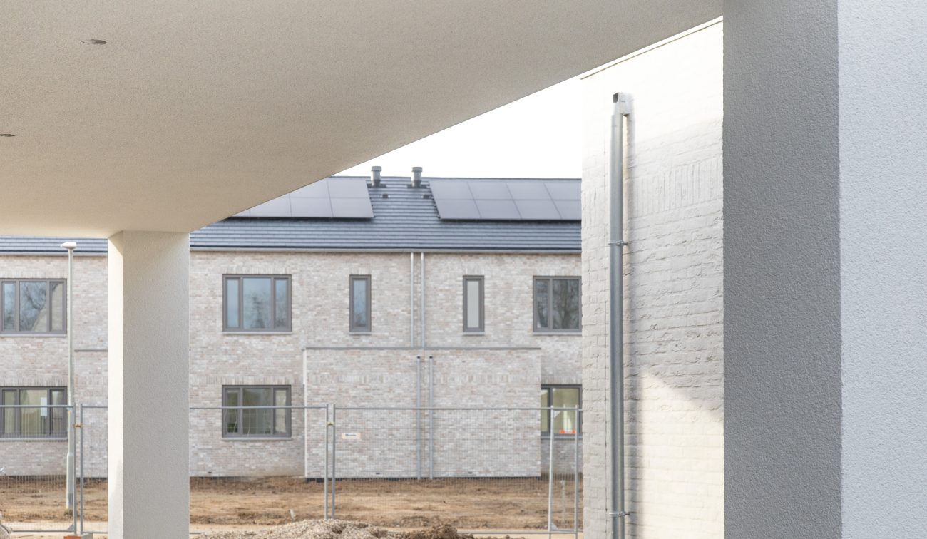 CPO Meerdel Elsloo – doorkijk onder wit gestuct overstek naar lichte bakstenen rijwoningen met zonnepanelen en bouwhek