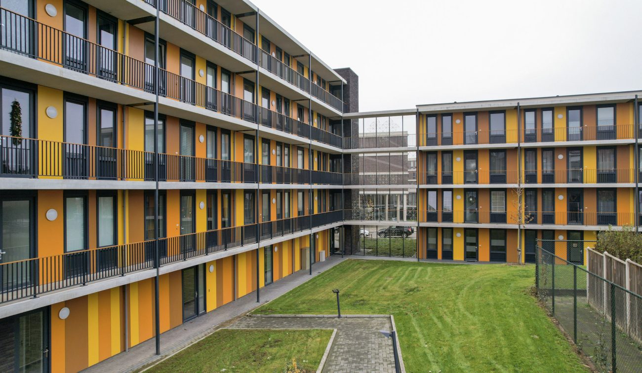 Uitzicht op het binnenterrein van een L-vormig appartementencomplex; ritmische gele en oranje gevelstroken, galerijen met zwarte hekwerken en een verzorgd grasveld.