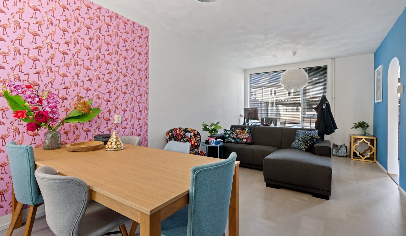 Woonkamer met roze flamingobehang, houten eettafel, grijze hoekbank, groot raam en blauwe accentwand
