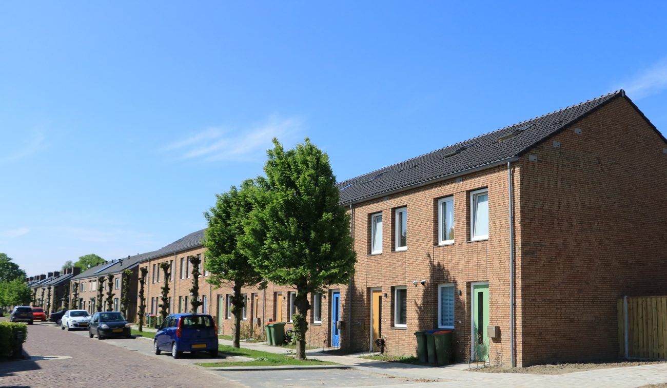 Conceptwoningen Klundert: rijwoningen in bakstenen met kleurige deuren, bomen en geparkeerde auto's.