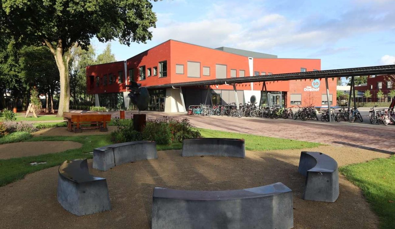 Nieuwbouw basisschool Den Doelhof in Meijel met rood gevelvolume en luifel; fietsenstalling met veel fietsen langs de gevel. Op de voorgrond een ronde zitplek van betonnen banken in de groene schooltuin met bomen.