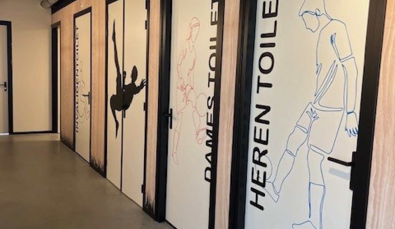 Interieur FC Landgraaf: corridor met grafische sportfiguren op wc-deuren en zichtbare techniek