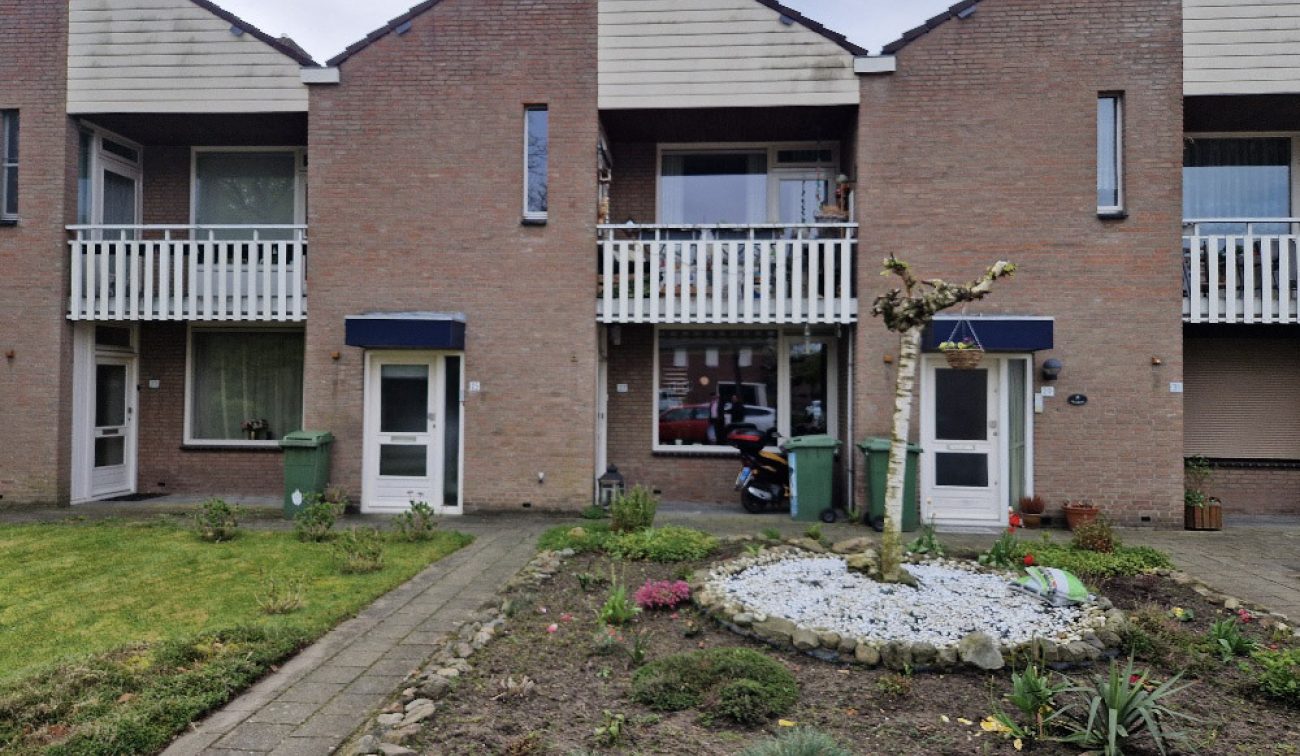 Portiekwoningen in Stramproy (Groot onderhoud 28 woningen) met bakstenen gevel, witte balkonhekken en blauwe luifels; twee entrees met stoep en groencontainers, voortuin met grindcirkel en gesnoeide boom, scooter achter raam zichtbaar.