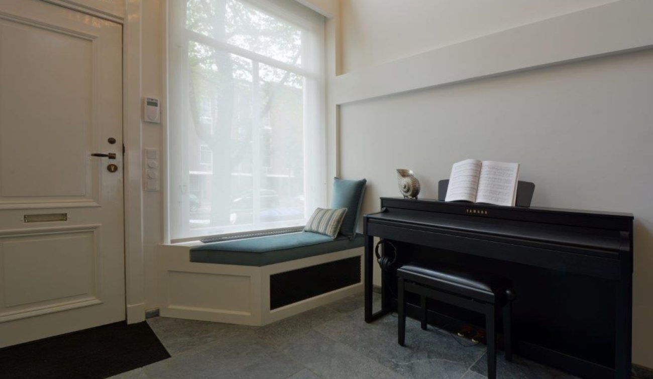 Entree met raamzitje, lichtfilter gordijn en zwarte piano; rustige, stijlvolle renovatie aan gracht