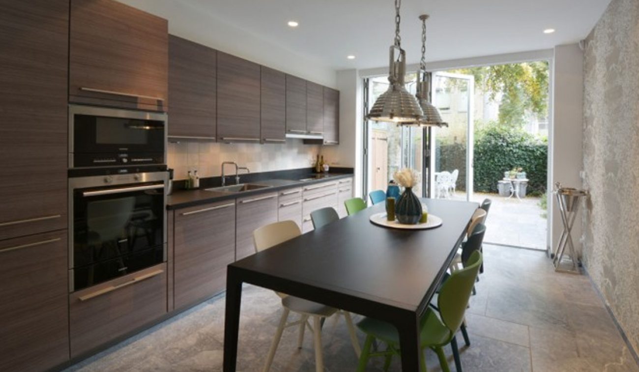 Renovatie Egelantiersgracht: moderne keukenwand, hanglampen boven tafel en zicht op patio met groen