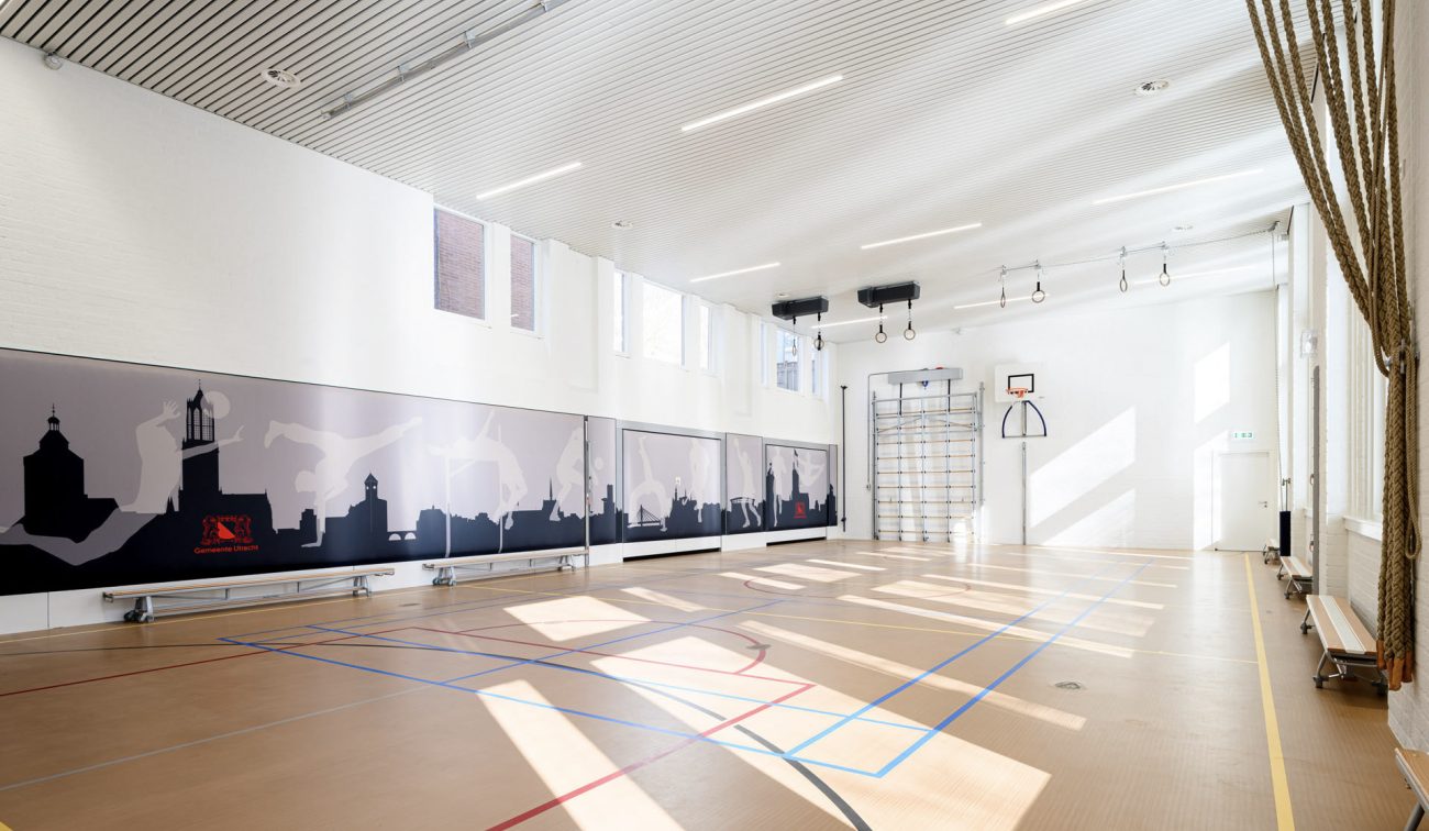 Renovatie gymzaal – hoge, lichte sportruimte met nieuwe sportvloer en belijning, akoestisch lamellenplafond, klimvoorzieningen (ringen/rek) en wandbescherming met grafische prints.
