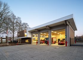 Nieuwe brandweerpost Sittard-Oost bij schemering met drie open uitrijpoorten; binnen staan een brandweerwagen en dienstauto klaar, naast de glazen entree in het moderne hout-wit gebouw.